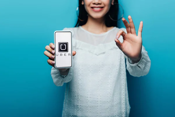 KYIV, UCRANIA - 15 de julio de 2019: vista recortada de una mujer asiática sonriente en blusa blanca que muestra un signo aceptable y un teléfono inteligente con aplicación uber sobre fondo azul - foto de stock