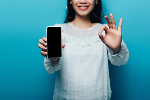 Обрезанный вид улыбающейся азиатской женщины в белой блузке показывает знак ОК и смартфон с чистым экраном на синем фоне — стоковое фото