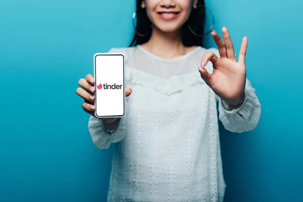 КИЕВ, УКРАИНА - 15 июля 2019 года: обрезанный вид улыбающейся азиатской женщины в белой блузке, показывающий знак ОК и смартфон с приложением TINder на синем фоне — стоковое фото