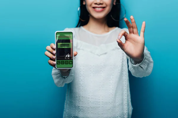 Recortado vista de sonriente mujer asiática en blusa blanca mostrando ok signo y teléfono inteligente con aplicación de reserva sobre fondo azul - foto de stock