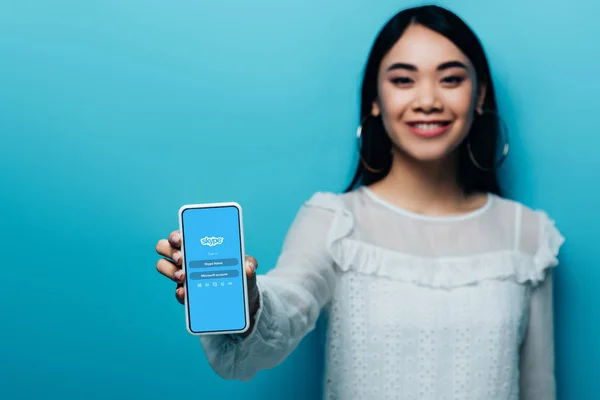 KYIV, UCRANIA - 15 de julio de 2019: enfoque selectivo de la sonriente mujer asiática en blusa blanca sosteniendo teléfono inteligente con aplicación skype sobre fondo azul - foto de stock