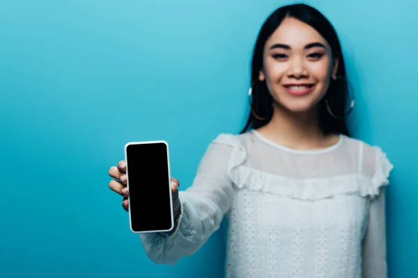 Messa a fuoco selettiva di donna asiatica sorridente in camicetta bianca che tiene smartphone con schermo bianco su sfondo blu — Foto stock