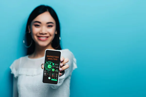 Селективный фокус улыбающейся азиатской женщины в белой блузке, держащей смартфон с приложением маркетингового анализа на синем фоне — стоковое фото