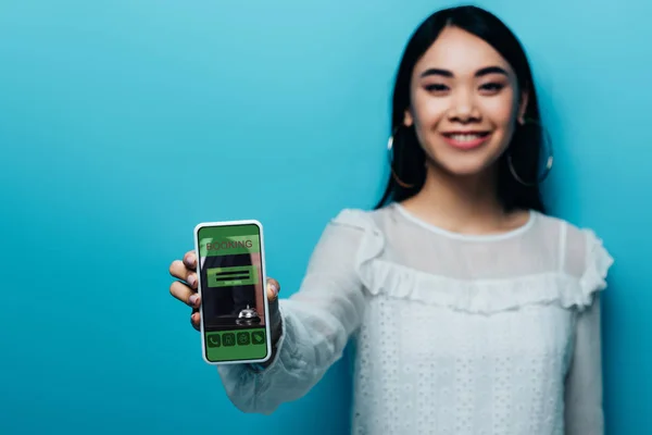 Селективный фокус улыбающейся азиатской женщины в белой блузке, держащей смартфон с приложением бронирования на синем фоне — стоковое фото