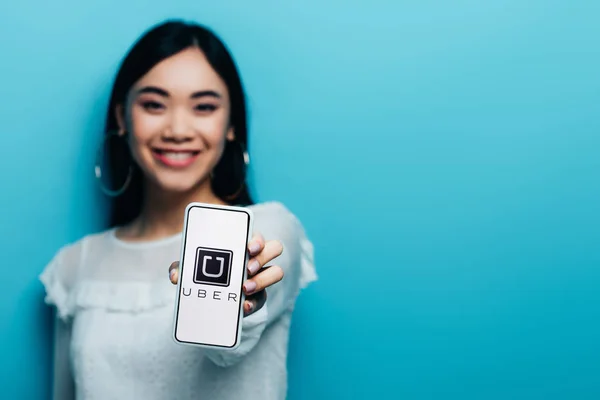 KYIV, UCRANIA - 15 de julio de 2019: enfoque selectivo de la sonriente mujer asiática en blusa blanca sosteniendo teléfono inteligente con aplicación uber sobre fondo azul - foto de stock