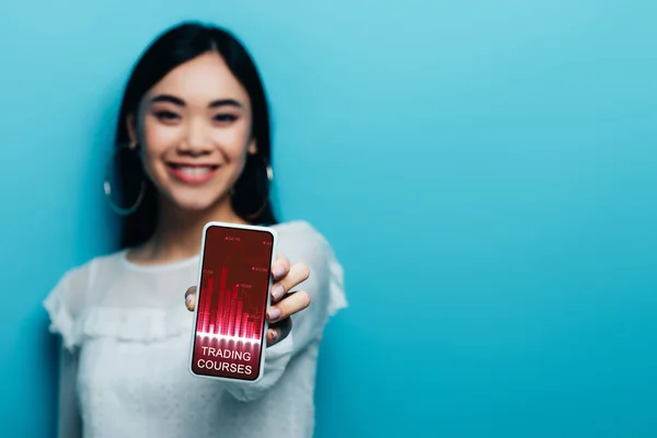 Enfoque selectivo de sonriente mujer asiática en blusa blanca celebración de teléfono inteligente con cursos de comercio aplicación sobre fondo azul - foto de stock