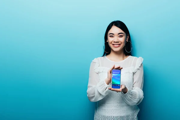 Усміхнена азіатська жінка в білій блузці тримає смартфон із додатком бронювання на синьому фоні — стокове фото