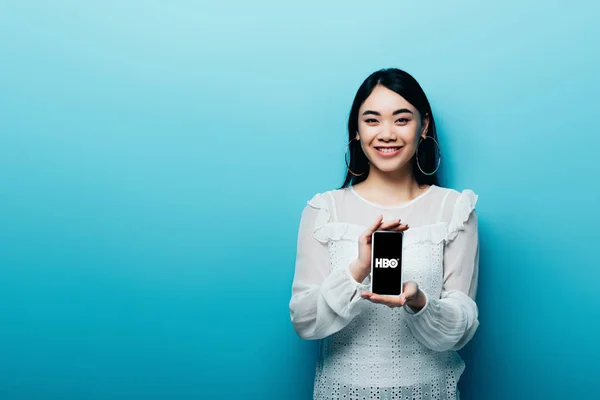 КИЕВ, Украина - 15 июля 2019 года: улыбающаяся азиатка в белой блузке держит смартфон со скайпом HBO на синем фоне — стоковое фото