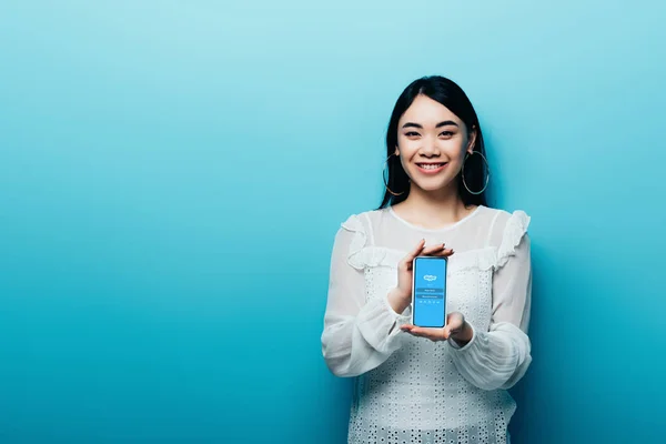 KYIV, UCRANIA - 15 de julio de 2019: mujer asiática sonriente en blusa blanca sosteniendo teléfono inteligente con aplicación skype sobre fondo azul - foto de stock