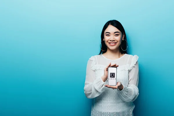 KYIV, UKRAINE - 15 JUILLET 2019 : femme asiatique souriante en chemisier blanc tenant smartphone avec application uber sur fond bleu — Photo de stock