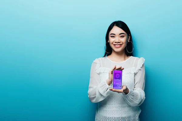 Улыбающаяся азиатка в белой блузке, держащая смартфон с приложением для покупок в интернете на синем фоне — стоковое фото