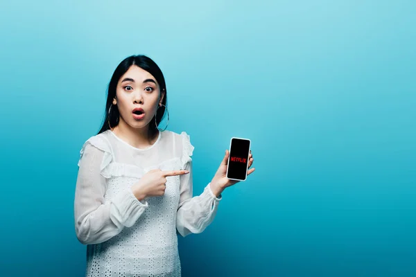 KYIV, UCRANIA - 15 de julio de 2019: mujer asiática impactada señalando con el dedo al teléfono inteligente con la aplicación netflix sobre fondo azul — Stock Photo