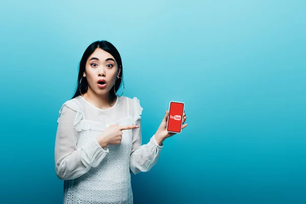 КИЕВ, Украина - 15 июля 2019 года: шокированная азиатская женщина, указывающая пальцем на смартфон с приложением youtube на синем фоне — стоковое фото