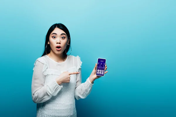 Conmocionado mujer asiática señalando con el dedo en el teléfono inteligente con aplicación sanitaria sobre fondo azul - foto de stock