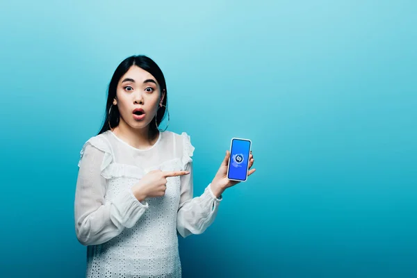 KYIV, UCRANIA - 15 de julio de 2019: mujer asiática impactada señalando con el dedo al teléfono inteligente con la aplicación Shazam sobre fondo azul - foto de stock