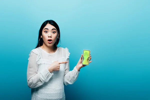 Shocked asian woman pointing with finger at smartphone with fashion shopping app on blue background — Stock Photo