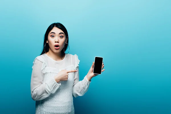 Chocado asiático mulher apontando com dedo para smartphone com tela em branco no fundo azul — Fotografia de Stock