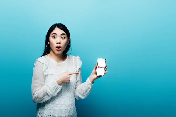 KYIV, UKRAINE - JULY 15, 2019: shocked asian woman pointing with finger at smartphone with tinder app on blue background — Stock Photo