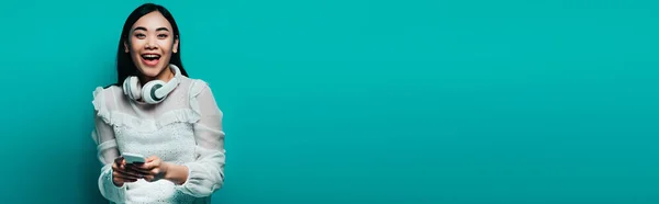 Панорамний знімок щасливої азіатки з бездротовими навушниками у білій блузці з використанням смартфона на бірюзовому фоні — стокове фото