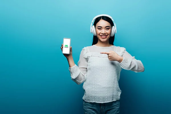 KYIV, UCRANIA - 15 de julio de 2019: mujer asiática sonriente con auriculares apuntando con el dedo al teléfono inteligente con el icono de Android sobre fondo azul - foto de stock