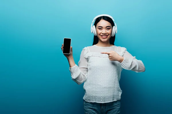 Усміхнена азіатська жінка з навушниками, спрямованими пальцем на смартфон з порожнім екраном на синьому фоні — стокове фото