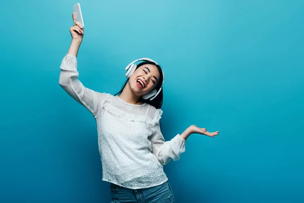 Улыбающаяся азиатка с закрытыми глазами в наушниках, держащая смартфон и танцующая на синем фоне — стоковое фото