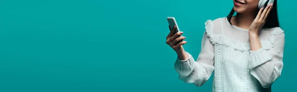Cropped view of smiling asian woman with headphones using smartphone on turquoise background, panoramic shot — Stock Photo