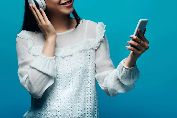 Abgeschnittene Ansicht einer lächelnden asiatischen Frau mit Kopfhörern mittels Smartphone auf blauem Hintergrund — Stockfoto