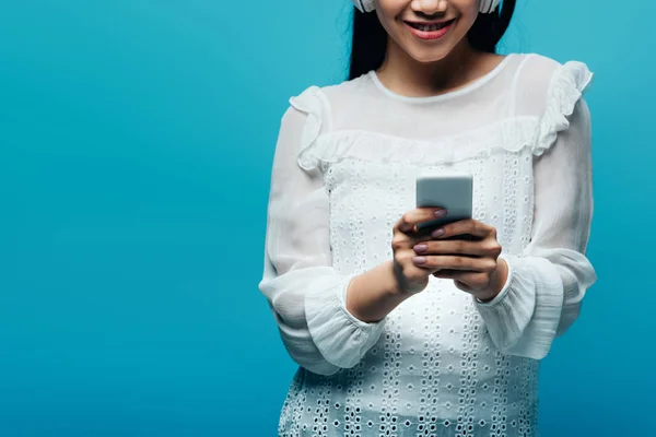 Vue partielle de souriant asiatique femme avec casque en utilisant smartphone sur fond bleu — Photo de stock