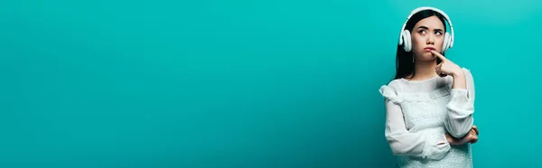 Мечтательная азиатка с наушниками, смотрящая на бирюзовый фон, панорамный снимок — стоковое фото