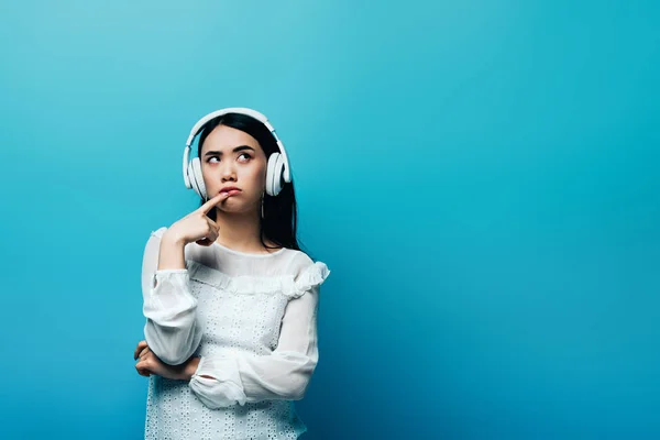 Verträumte asiatische Frau mit Kopfhörern auf blauem Hintergrund — Stockfoto