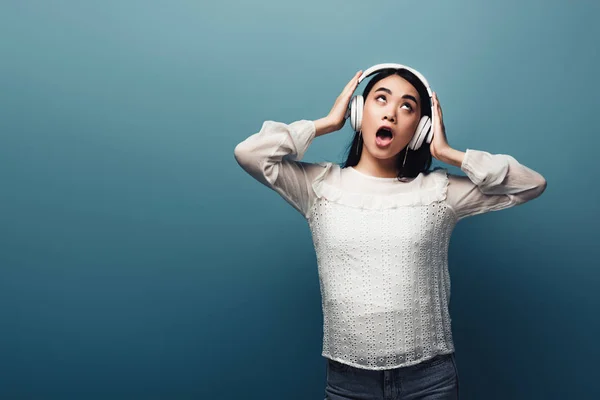 Impactado mujer asiática con las manos en los auriculares y la boca abierta sobre fondo azul - foto de stock