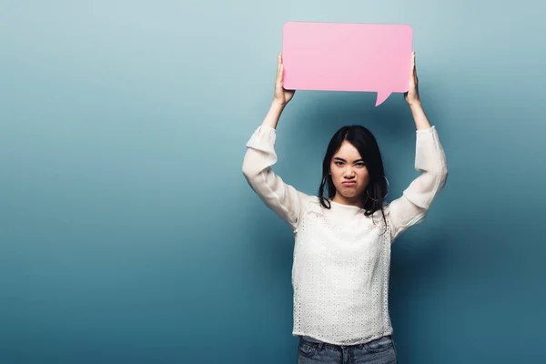 Displeased brunette asian woman holding pink speech bubble on blue background — Stock Photo