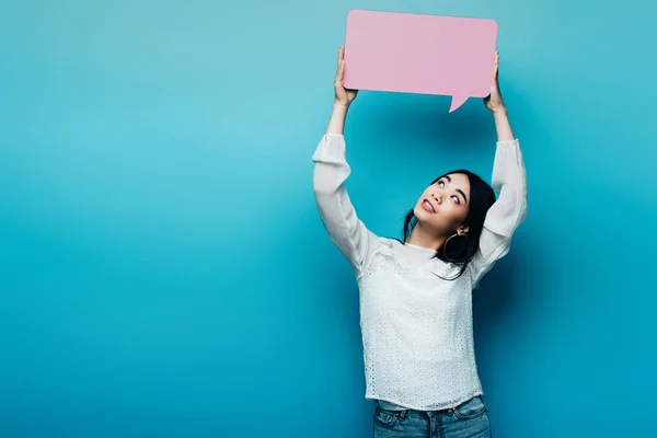 Smiling brunette asian woman holding pink speech bubble on blue background — Stock Photo