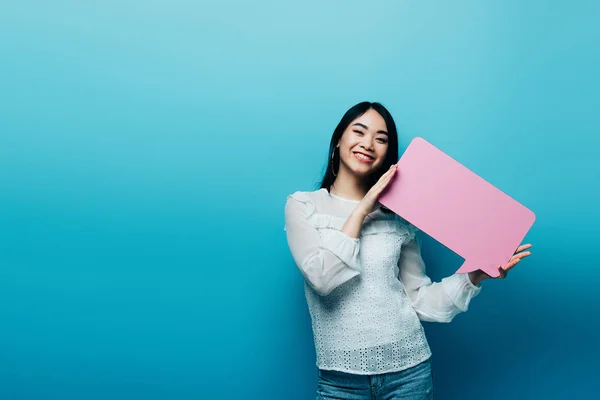 Cheerful brunette asian woman holding pink speech bubble on blue background — Stock Photo