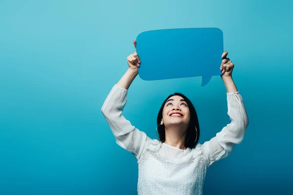 Feliz morena asiático mujer mirando discurso burbuja en azul fondo - foto de stock