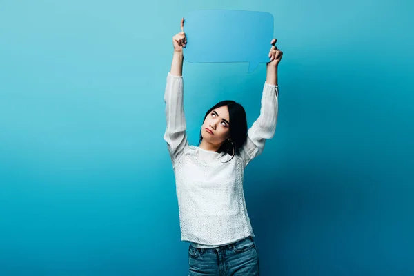 Засмучена брюнетка азіатська жінка тримає мовну бульбашку на синьому фоні — стокове фото