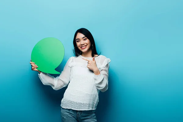 Lächelnde brünette asiatische Frau zeigt mit dem Finger auf grüne Sprechblase auf blauem Hintergrund — Stockfoto