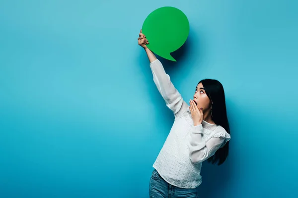 Shocked brunette asian woman holding green speech bubble on blue background — Stock Photo