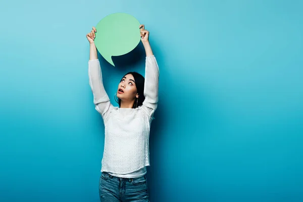 Surprised brunette asian woman looking at green speech bubble on blue background — Stock Photo