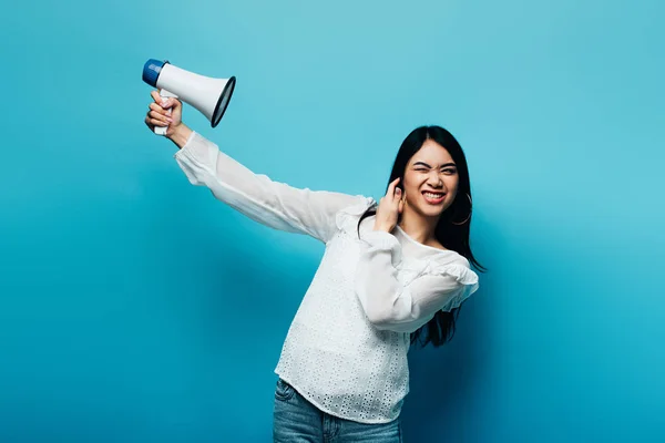 Brunette asian woman holding loudspeaker on blue background — Stock Photo