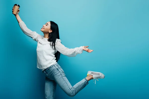 Happy brunette asian woman posing with coffee to go on blue background — Stock Photo