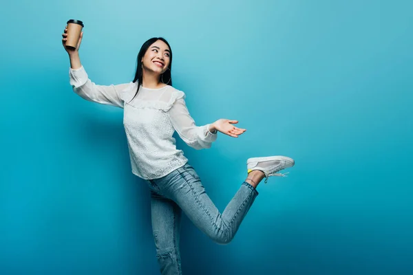 Happy brunette asian woman posing with coffee to go on blue background — Stock Photo
