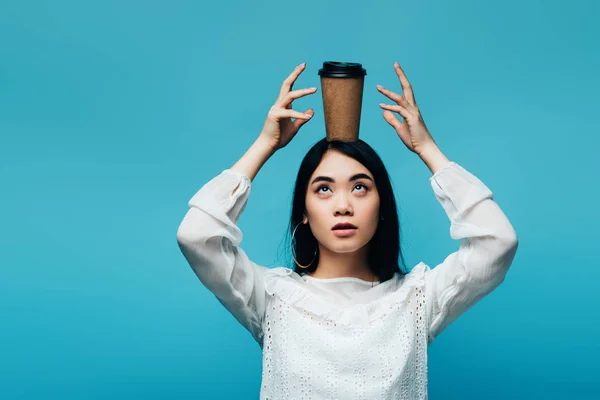 Brunette asian woman putting coffee to go on head on blue background — Stock Photo