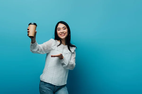Smiling brunette asian woman pointing with hand at coffee to go on blue background — Stock Photo