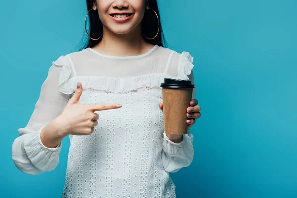 Abgeschnittene Ansicht der lächelnden brünetten asiatischen Frau, die mit dem Finger auf Kaffee zeigt, um auf blauem Hintergrund zu gehen — Stockfoto