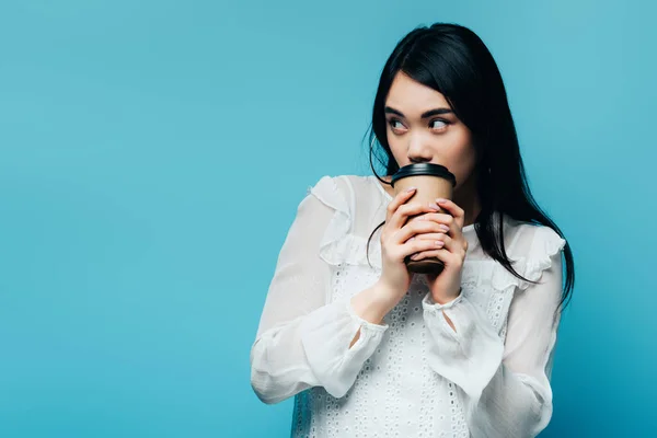 Brunette asiatique femme tenant café pour aller et regarder loin sur fond bleu — Photo de stock