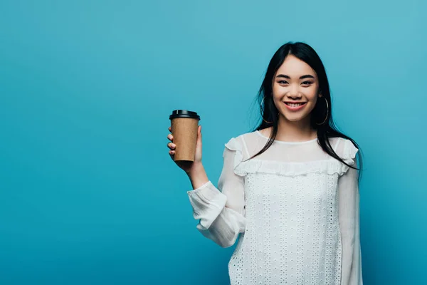 Smiling brunette asian woman holding coffee to go on blue background — Stock Photo