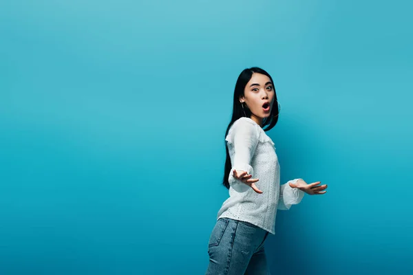 Excited brunette asian woman gesturing with open mouth on blue background — Stock Photo