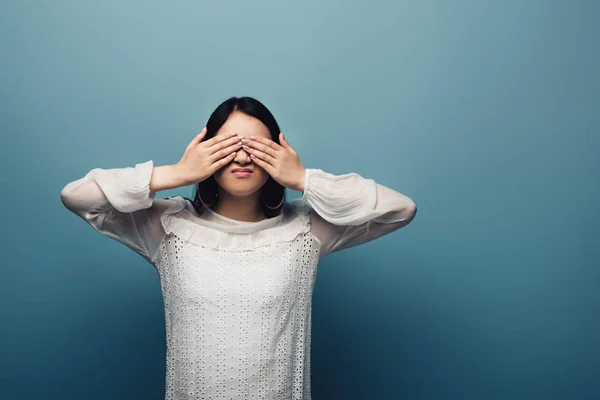 Засмучена брюнетка азіатська жінка з руками на очах на синьому фоні — стокове фото